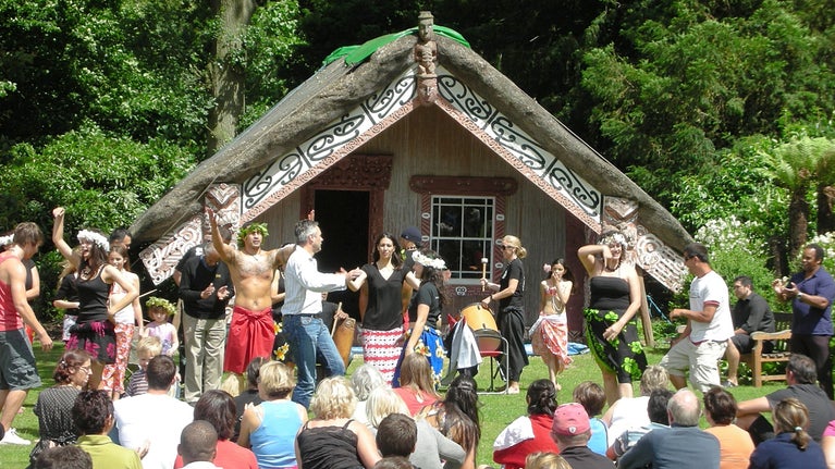 A group of people performing traditional Māori dance in front of a Māori meeting house with an audience sitting on the ground watching